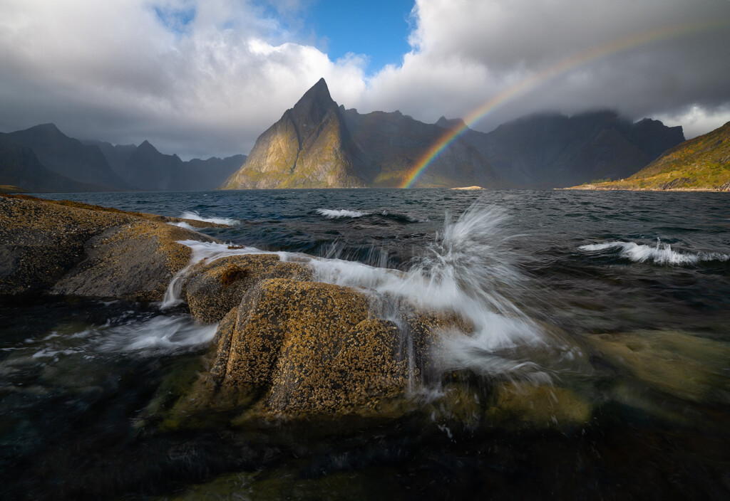 Waves crash against the shoreline as sunlight spills through small openings in the clouds to illuminate one of Norways most iconic peaks in Hamnøy